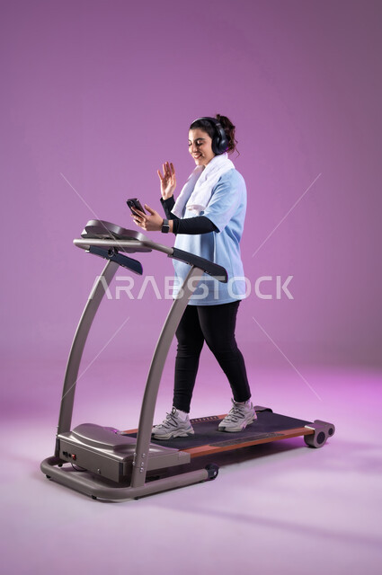 قضاء وقت ممتع أثناء التمرين على جهازالمشي، القاء التحية والسلام، استخدام الأجهزة التقنية الحديثة، مفهوم الرشاقة واللياقة، بورتريه لإمرأة عربية سعودية خليجية ترتدي الملابس الرياضية تضع الهيدفون وتجري مكالمة فيديوعبر الهاتف المحمول، الطول الكامل للجسم، خلفية بنفسجية