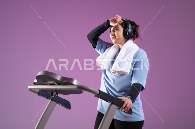 الشعور بالتعب والارهاق اثناء التمرين, التدرب بعزيمة للمحافظة على نظام الحياة الصحي، انشطة شبابية تساعد على اللياقة البدنية, اهمية الرياضة لبناء جسد قوي, بورتريه مقرب لفتاة عربية خليجية سعودية ترتدي الملابس الرياضية وتضع سماعة الهيدفون ، خلفية بنفسجية