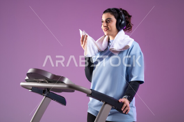 التدرب بعزيمة للمحافظة على نظام الحياة الصحي، انشطة شبابية تساعد على اللياقة البدنية, اهمية الرياضة لبناء جسد قوي, بورتريه مقرب لفتاة عربية خليجية سعودية ترتدي الملابس الرياضية وتضع سماعة الهيدفون تستخدم المنشفة لتجفيف العرق أثناء التمرين، خلفية بنفسجية