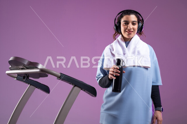 جودة الحياة الصحية، نشاط بدني للمحافظة على اللياقة الجسدية، بورتريه مقرب لشابة عربية خليجية سعودية مبتسمة ترتدي الملابس الرياضية وتضع الهيدفون تقوم بشرب الماء أثناء التدريب على العجلة الهوائية، دراجة تمارين ثابتة، خلفية بنفسجية