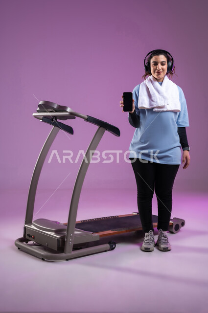 ممارسة الهوايات المفيدة، عرض شاشة سوداء فارغة على الهاتف المحمول، قضاء وقت ممتع على العجلة الهوائية، دراجة تمارين ثابتة، بورتريه لفتاة عربية سعودية خليجية ترتدي الملابس الرياضية وتضع الهيدفون، المحافظة على نظام الحياة الصحي، الطول الكامل للجسم، خلفية بنفسجية