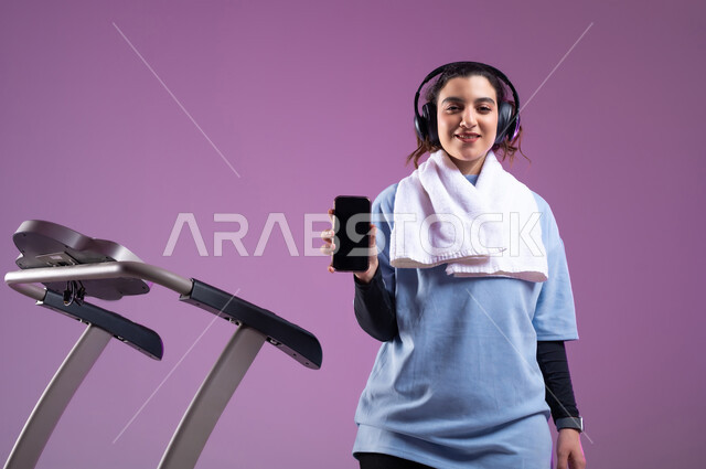 عرض شاشة سوداء فارغة على الهاتف المحمول، ممارسة الهوايات المفيدة،  قضاء وقت ممتع على العجلة الهوائية، دراجة تمارين ثابتة، بورتريه مقرب لفتاة عربية سعودية خليجية ترتدي الملابس الرياضية وتضع الهيدفون، المحافظة على نظام الحياة الصحي، خلفية بنفسجية