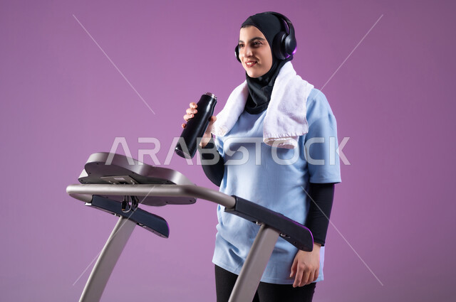 جودة الحياة الصحية، نشاط بدني للمحافظة على اللياقة الجسدية، بورتريه لشابة عربية خليجية سعودية مبتسمة ترتدي الملابس الرياضية وتضع الهيدفون تقوم بشرب الماء أثناء التدريب على العجلة الهوائية، دراجة تمارين ثابتة، الطول الكامل للجسم، خلفية بنفسجية
