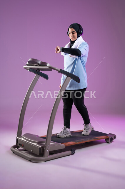 التدرب بعزيمة للمحافظة على نظام الحياة الصحي،ايماءات السعادة والنشاط، مفهوم الرشاقة واللياقة، بورتريه لفتاة عربية خليجية سعودية ترتدي ملابس الرياضية تضع الهيدفون تتابع تقدمها في الأداء على جهاز المشي، ممارسة الهوايات المفيدة، خلفية بنفسجية