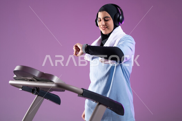 ايماءات السعادة والنشاط، مفهوم الرشاقة واللياقة، بورتريه مقرب لفتاة عربية خليجية سعودية ترتدي الملابس الرياضية تضع الهيدفون على رأسها تتتابع تقدمها في الأداء على جهاز المشي، ممارسة الهوايات المفيدة، التدرب بعزيمة للمحافظة على نظام الحياة الصحي، خلفية بنفسجية
