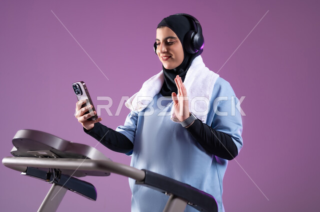 Making a video call through social networking sites, a healthy lifestyle, physical activity to maintain health and physical fitness, a close-up portrait of a veiled Saudi Gulf Arab girl wearing clothes of an athlete with a headphone on her head doing warm-up exercises on a treadmill, purple background