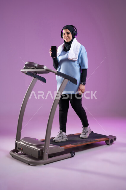 نشاط بدني للمحافظه على الصحه واللياقه الجسديه, بورتريه لشابة عربية خليجية سعودية محجبة ترتدي الملابس الرياضية تضع على رأسها سماعة الهيدفون, القيام بتمارين الإحماء على جهاز المشي، عرض شاشة سوداء فارغة على الهاتف المحمول، أسلوب حياة صحي، الطول الكامل للجسم، خلفية بنفسجية