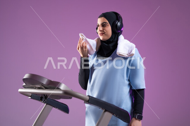 المحافظة على نظام الحياة الصحي، انشطة شبابية تساعد على اللياقة البدنية, اهمية الرياضة لبناء جسد قوي, بورتريه مقرب لشابة عربية خليجية سعودية محجبة ترتدي الملابس الرياضية وتضع سماعة الهيدفون, استخدام المنشفة لتجفيف العرق أثناء التمرين، خلفية بنفسجية
