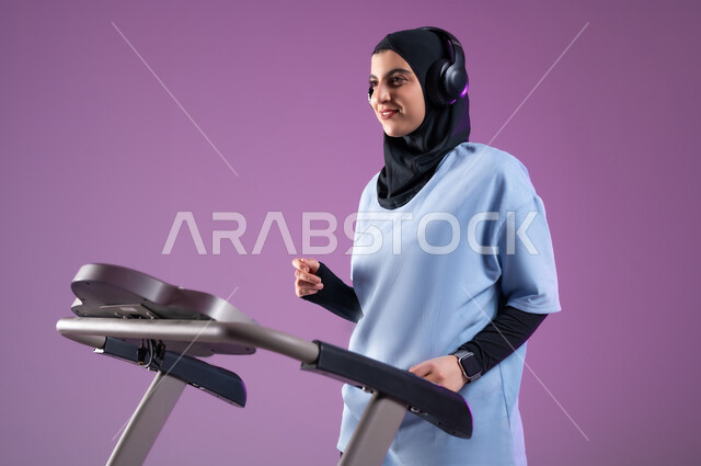 تمارين شبابية تساعد على اللياقة البدنية, المحافظة على نظام الحياة الصحي لبناء جسد قوي, بورتريه مقرب لشابة عربية خليجية سعودية محجبة مبتسمة ترتدي الملابس الرياضية و تضع على رأسها سماعة الهيدفون, الاحماء على جهاز المشي، خلفية بنفسجية