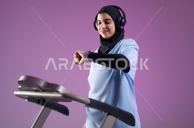 أنشطة شبابية تساعد على اللياقة البدنية، المحافظة على نظام الحياة الصحي، بورتريه لشابة عربية خليجية سعودية محجبة ترتدي الملابس الرياضية تضع على رأسها سماعة الهيدفون, متابعة التقدم في الأداء على جهاز المشي،  خلفية بنفسجية