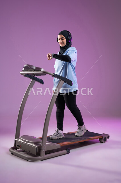 التدرب بجد وعزيمة للمحافظة على نظام الحياة الصحي، بورتريه لشابة عربية خليجية سعودية محجبة ترتدي الملابس الرياضية تضع على رأسها سماعة الهيدفون, متابعة التقدم  في الأداء على جهاز المشي،  أنشطة شبابية تساعد على اللياقة البدنية, صورة الجسم بالطول الكامل، خلفية بنفسجية
