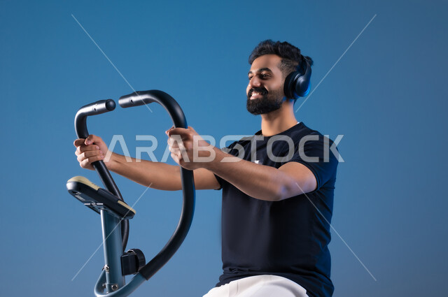 Maintaining a healthy lifestyle, youth activities that help with physical fitness, a close-up portrait of a smiling Saudi Arabian Gulf young man wearing sports clothes and wearing a headphone, training on the sports equipment with enthusiasm, blue background