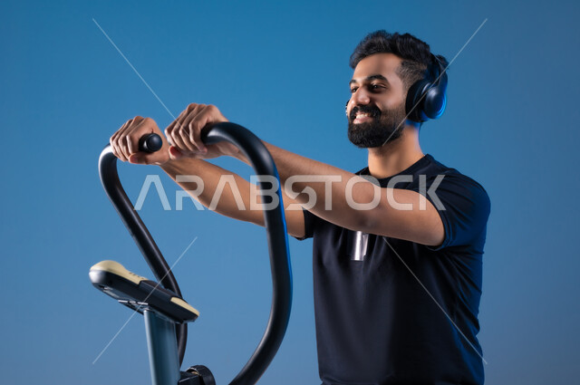 The importance of exercise to build a strong body, a close-up portrait of a smiling Saudi Gulf Arab young man wearing sports clothes and wearing a headphone, training on the sports equipment with enthusiasm, maintaining a healthy lifestyle, youth activities that help with physical fitness, blue background