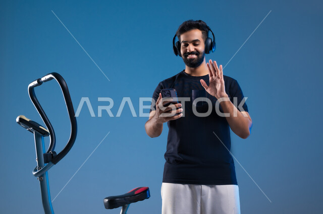 استخدام الأجهزة التقنية الحديثة، مفهوم الرشاقة واللياقة، أسلوب حياة صحية، بورتريه لشاب عربي سعودي خليجي يرتدي الملابس الرياضية على رأسه سماعة الهيدفون, القيام بإجراء مكالمة فيديوعبر الهاتف المحمول،القاء التحية والسلام، قضاء وقت ممتع أثناء التمرين،خلفية زرقاء