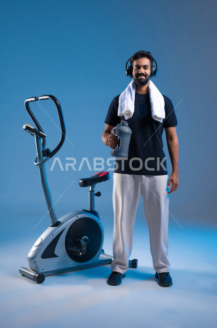 نشاط بدني للحفاظ على الصحة الجيدة, اهمية شرب الماء أثناء التمرين لبناء جسد قوي، بورتريه لشاب عربي سعودي خليجي مبتسم يرتدي الملابس الرياضية يضع على رأسه سماعة الهيدفون, مفهوم الراقة واللياقة، صورة الجسم بالطول الكامل، خلفية زرقاء