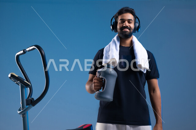 اهمية شرب الماء أثناء التمرين لبناء جسد قوي، بورتريه مقرب لشاب عربي سعودي خليجي مبتسم يرتدي الملابس الرياضية يضع سماعة الهيدفون يقوم بالتدريب على العجلة الرياضية، المحافظة على نظام الحياة الصحي, أنشطة شبابية تساعد على اللياقة البدنية, خلفية زرقاء