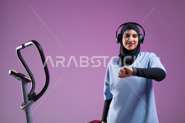 مفهوم الرشاقة واللياقة، بورتريه لشابة عربية خليجية سعودية محجبة ترتدي الملابس الرياضية تضع على رأسها سماعة الهيدفون, مراقبة وقت التمرين على العجلة الهوائية، ممارسة الهوايات المفضلة، دراجة تمارين ثابتة, المحافظة على نظام الحياة الصحي، خلفية بنفسجية