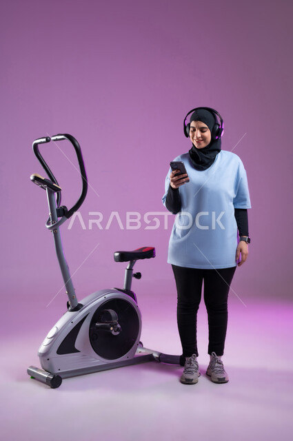 استخدام الأجهزة التقنية الحديثة، مفهوم الرشاقة واللياقة، بورتريه لشابة عربية خليجية محجبة سعودية ترتدي الملابس الرياضية تضع على رأسها سماعة الهيدفون, الاستعداد لبدء التمين على جهاز العجلة الهوائية, اختيار الموسيقى المفضلة عبر الجوال، دراجة التمارين الثابتة, الطول الكامل للجسم،  خلفية بنفسجية