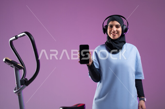 دراجة تمارين ثابتة, عرض شاشة سوداء فارغة على الهاتف المحمول، ممارسة الهوايات المفضلة، قضاء وقت ممتع على العجلة الهوائية، بورتريه لشابة عربية سعودية خليجية محجبة ترتدي الملابس الرياضية وتضع على رأسها سماعة الهيدفون، المحافظة على اللياقة والرشاقة البدنية، أسلوب حياة صحي, خلفية بنفسجية