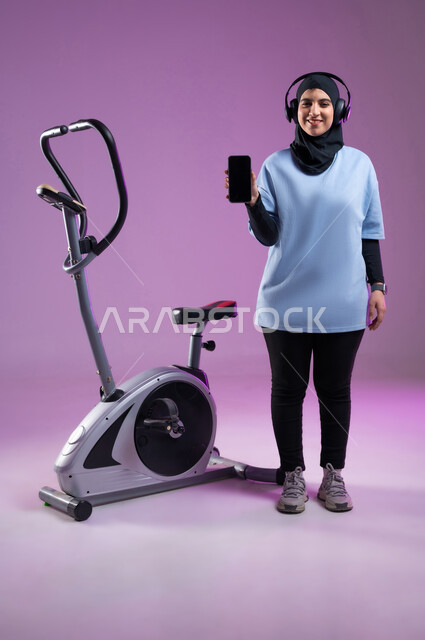 ممارسة الهوايات المفضلة، عرض شاشة سوداء فارغة على الهاتف المحمول،  قضاء وقت ممتع على العجلة الهوائية، بورتريه لشابة عربية سعودية خليجية محجبة ترتدي الملابس الرياضية وتضع على رأسها سماعة الهيدفون, المحافظة على الرشاقة واللياقة البدنية، الطول الكامل للجسم، خلفية بنفسجية