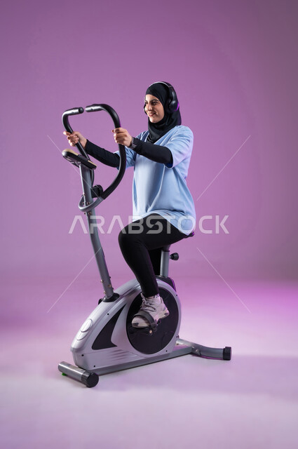 Maintaining a healthy lifestyle, youth activities that help with physical fitness, a portrait of a veiled Saudi Arabian Gulf girl, smiling, wearing sports clothes and wearing a headphone, training on the sports equipment with enthusiasm, full body length, colorful background