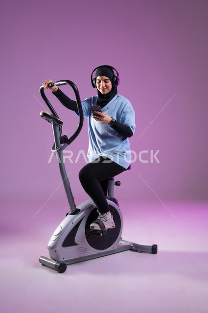 استخدام الأجهزة التقنية الحديثة، بورتريه لشابة عربية خليجية سعودية ترتدي الملابس  الرياضية تضع على رأسها سماعة الهيدفون, دراجة تمارين ثابتة, الاستماع للموسيقى أثناء ممارسة التمارين الرياضية على جهاز العجلة الهوائية، مفهوم حرق الدهون و إنقاص الوزن، الطول الكامل للجسم، خلفية بنفسجية