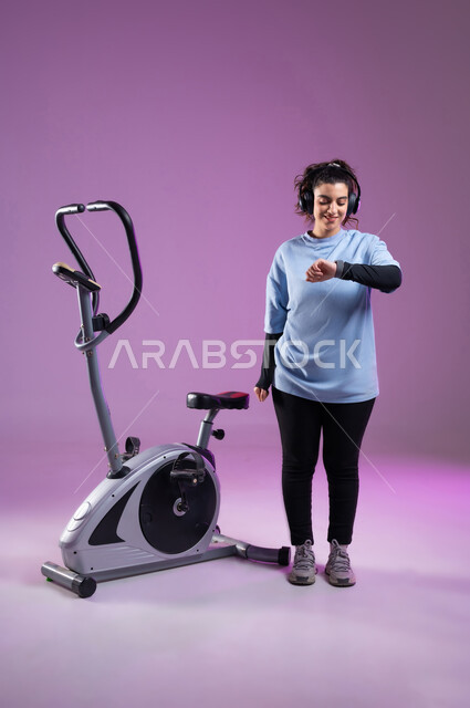 مفهوم الرشاقة واللياقة البدنية، بورتريه لشابة عربية خليجية سعودية ترتدي الملابس الرياضية تضع على رأسها سماعة الهيدفون, ممارسة الهوايات المفضلة،  مراقبة الوقت لبدء التمرين على العجلة الهوائية، دراجة تمارين ثابتة, المحافظة على نظام الحياة الصحي، الطول الكامل للجسم، خلفية بنفسجية