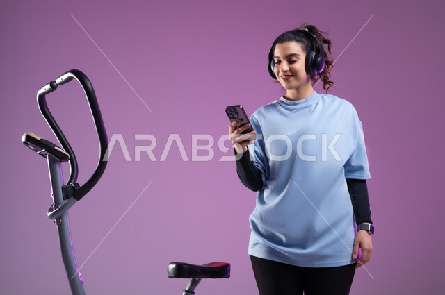 استخدام الأجهزة التقنية الحديثة، مفهوم الرشاقة واللياقة، بورتريه لشابة عربية خليجية سعودية ترتدي الملابس الرياضية تضع على رأسها سماعة الهيدفون, الاستعداد لبدء التمين على جهاز العجلة الهوائية, اختيار الموسيقى المفضلة عبر الجوال، دراجة التمارين الثابتة,  خلفية بنفسجية