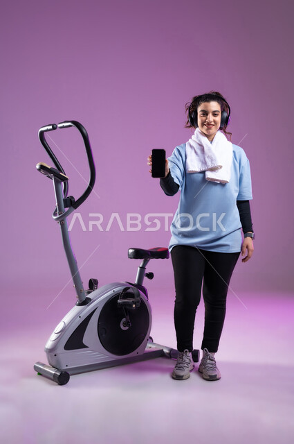 ممارسة الهوايات المفضلة، عرض شاشة سوداء فارغة على الهاتف المحمول،  قضاء وقت ممتع على العجلة الهوائية، بورتريه لشابة عربية سعودية خليجية ترتدي الملابس الرياضية وتضع على رأسها سماعة الهيدفون, المحافظة على الرشاقة واللياقة البدنية، الطول الكامل للجسم، خلفية بنفسجية