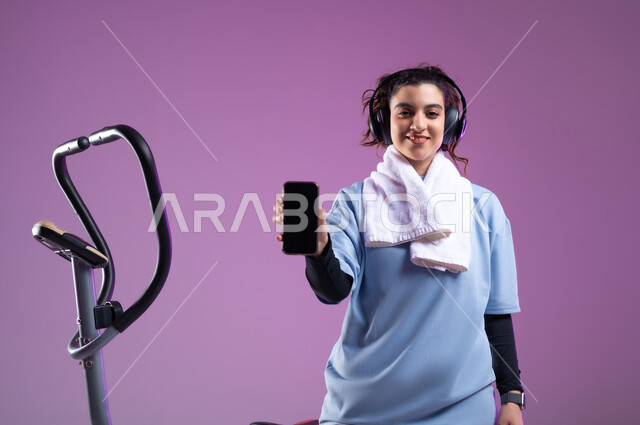 دراجة تمارين ثابتة, عرض شاشة سوداء فارغة على الهاتف المحمول، ممارسة الهوايات المفضلة، قضاء وقت ممتع على العجلة الهوائية، بورتريه لشابة عربية سعودية خليجية ترتدي الملابس الرياضية وتضع على رأسها سماعة الهيدفون، المحافظة على اللياقة والرشاقة البدنية، أسلوب حياة صحي, خلفية بنفسجية