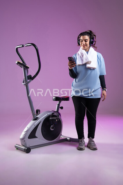 استخدام الأجهزة التقنية الحديثة، مفهوم الرشاقة واللياقة، بورتريه لشابة عربية خليجية سعودية ترتدي الملابس الرياضية تضع على رأسها سماعة الهيدفون, الاستعداد لبدء التمين على جهاز العجلة الهوائية, اختيار الموسيقى المفضلة عبر الجوال، دراجة التمارين الثابتة, الطول الكامل للجسم،  خلفية بنفسجية