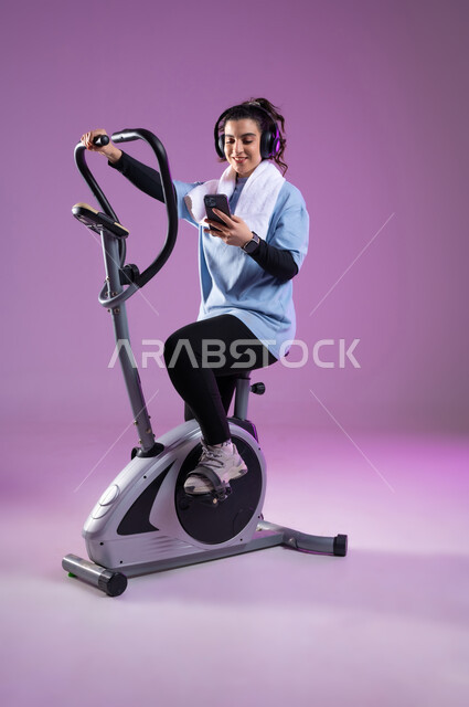 استخدام الأجهزة التقنية الحديثة، بورتريه لشابة عربية خليجية سعودية ترتدي الملابس  الرياضية تضع على رأسها سماعة الهيدفون, دراجة تمارين ثابتة, الاستماع للموسيقى أثناء ممارسة التمارين الرياضية على جهاز العجلة الهوائية، مفهوم حرق الدهون و إنقاص الوزن، الطول الكامل للجسم، خلفية بنفسجية