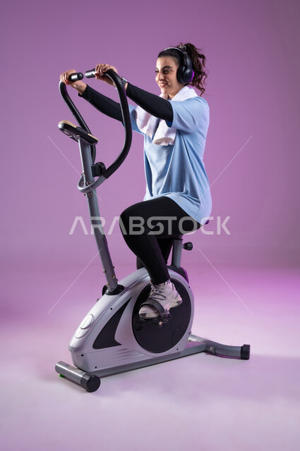 Maintaining a healthy lifestyle, youth activities that help with physical fitness, a portrait of a smiling Saudi Arabian Gulf girl wearing sports clothes and wearing a headphone, training on the sports equipment with enthusiasm, full length of the body, colorful background