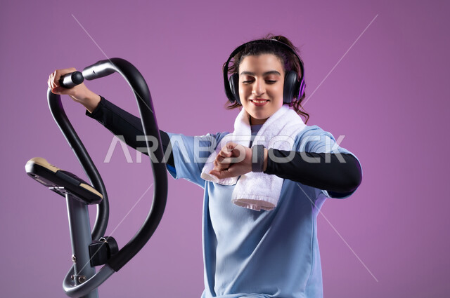 التدرب على جهاز العجلة الهوائية بتعابير السعادة والحماس، مفهوم الرشاقة واللياقة، بورتريه مقرب لفتاة عربية خليجية سعودية ترتدي الملابس الرياضية تضع على رأسها سماعة الهيدفون تتابع تقدمها في الأداء ، ممارسة الهوايات المفضلة، المحافظة على نظام الحياة الصحي، خلفية بنفسجية