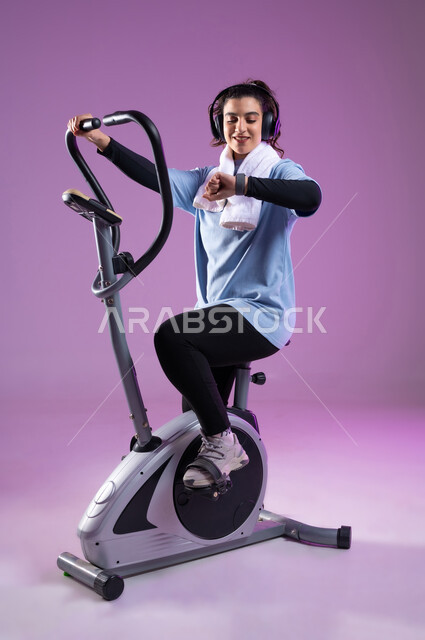 التدرب على جهاز العجلة الهوائية بتعابير الحماس والسعادة, المحافظة على نظام الحياة الصحي، بورتريه لفتاة عربية خليجية سعودية ترتدي الملابس الرياضية تضع على رأسها سماعة الهيدفون، دراجة تمارين ثابتة, أنشطة شبابية تساعد على اللياقة البدنية, صورة الجسم بالطول الكامل، خلفية بنفسجية