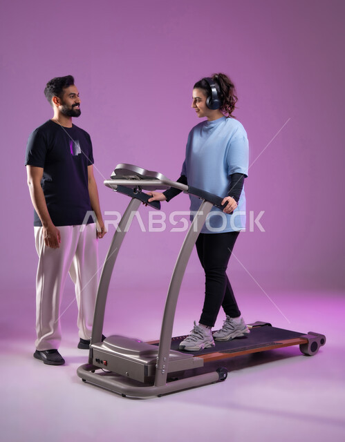 نشاط بدني للمحافظه على الصحه واللياقه الجسديه, مفهوم حرق الدهون و إنقاص الوزن، بورتريه لمدرب عربي خليجي سعودي يقوم بتدريب امرأة على تمارين الإحماء، الاستمتاع في قضاء أوقات ممتعة على جهاز العجلة الهوائية، أسلوب حياة صحي، الطول الكامل للجسم، خلفية بنفسجية