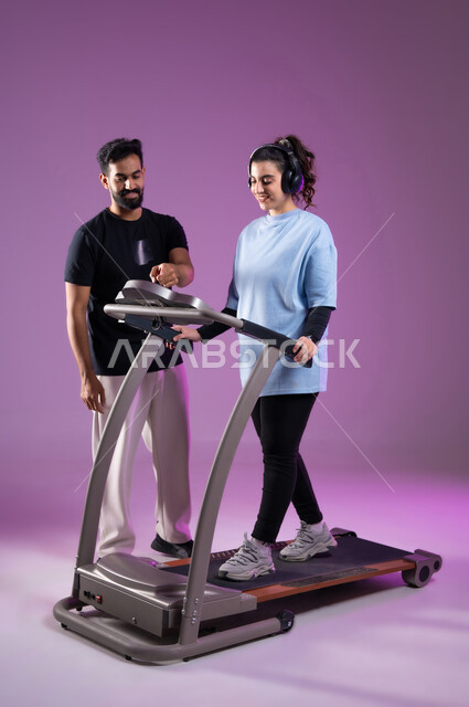 مفهوم حرق الدهون و إنقاص الوزن، نشاط بدني للمحافظه على الصحه واللياقه الجسديه, بورتريه لمدرب عربي خليجي سعودي يقوم بتدريب امرأة على تمارين الإحماء، الاستمتاع في قضاء أوقات ممتعة على جهاز العجلة الهوائية، أسلوب حياة صحي، الطول الكامل للجسم، خلفية بنفسجية