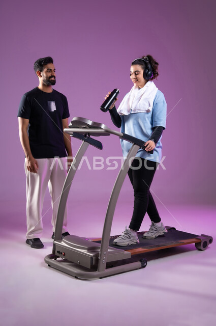 ممارسة الهوايات المفضلة، اهمية شرب الماء أثناء التمرين لبناء جسد قوي، بورتريه لمدرب عربي خليجي سعودي يرتدي الملابس الرياضية يقوم بتدريب امرأة على جهاز العجلة الهوائية، أسلوب حياة صحي، أنشطة شبابية تساعد على اللياقة البدنية, الطول الكامل للجسم، خلفية بنفسجية