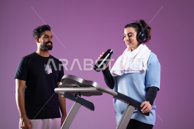 اهمية شرب الماء أثناء التمرين لبناء جسد قوي، بورتريه لمدرب عربي خليجي سعودي يرتدي الملابس الرياضية يقوم بتدريب امرأة على جهاز العجلة الهوائية، المحافظة على نظام الحياة الصحي, أنشطة شبابية تساعد على اللياقة البدنية, ممارسة الهوايات المضلة، خلفية بنفسجية