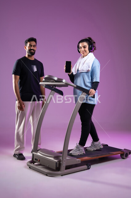 ممارسة الهوايات المفضلة، عرض شاشة سوداء فارغة على الهاتف المحمول، بورتريه  لمدرب عربي خليجي سعودي يرتدي الملابس الرياضية يقوم بتدريب امرأة على جهاز العجلة الهوائية، المحافظة على نظام الحياة الصحي، مفهوم الرشاقة واللياقة، الطول الكامل للجسم، خلفية بنفسجية