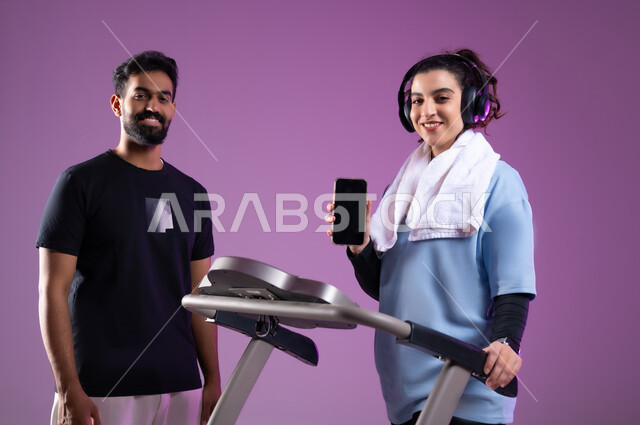 عرض شاشة سوداء فارغة على الهاتف المحمول، ممارسة الهوايات المفضلة، قضاء وقت ممتع أثناء التمرين، بورتريه لمدرب عربي خليجي سعودي يرتدي الملابس الرياضية يقوم بتدريب امرأة على جهاز العجلة الهوائية، المحافظة على نظام الحياة الصحي، مفهوم الرشاقة واللياقة، خلفية بنفسجية