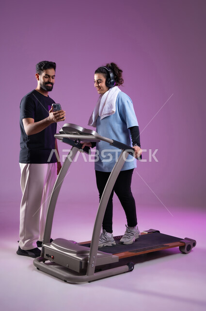 إجراء مكالمة فيديو مع الأصدقاء، المحافظة على نظام الحياة الصحي, بورتريه لمدرب عربي خليجي سعودي يرتدي الملابس الرياضية يقوم بتدريب امرأة على جهاز العجلة الهوائية، ايماءات السعادة والنشاط، استخدام الأجهزة التقنية الحديثة, الطول الكامل للجسم، خلفية بنفسجية