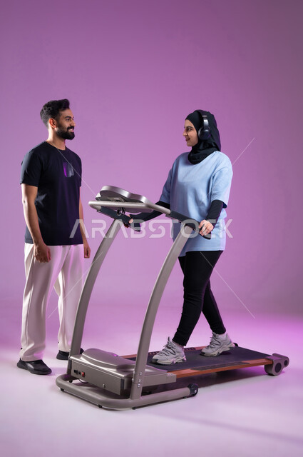 نشاط بدني للمحافظه على الصحه واللياقه الجسديه, مفهوم حرق الدهون و إنقاص الوزن، بورتريه لمدرب عربي خليجي سعودي يقوم بتدريب امرأة على تمارين الإحماء، الاستمتاع في قضاء أوقات ممتعة على جهاز العجلة الهوائية، دراجة تمارين ثابتة، الطول الكامل للجسم، خلفية بنفسجية