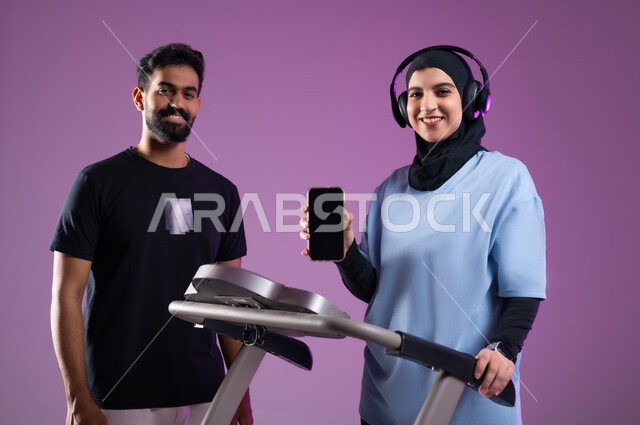 استخدام جهاز المشي لحرق الدهون، متدربة  تمسك بيدها الهاتف المحمول، عرض شاشة سوداء فارغة عبر جهاز الجوال، بورتريه لامراة  وشاب عربيان خليجيان سعوديان ، اسلوب حياة صحي، مفهوم الرشاقة واللياقة البدنية، المحافظة على التوازن الجسدي, خلفية بنفسجية
