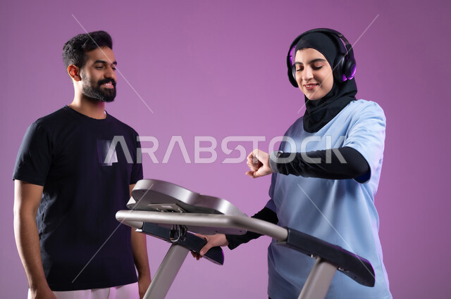 ارتداء ساعة باليد لتحكيم الوقت انثاء استخدام جهاز المشي لحرق الدهون، بورتريه مقرب لمدرب عربي خليجي سعودي يرتدي الملابس الرياضية يقوم بتدريب امرأة على جهاز العجلة الهوائية، دراجة التمرينات الثابتة، اسلوب حياة صحي، مفهوم الرشاقة واللياقة البدنية، انشطة ورياضات لتقوية العضلات, خلفية بنفسجية