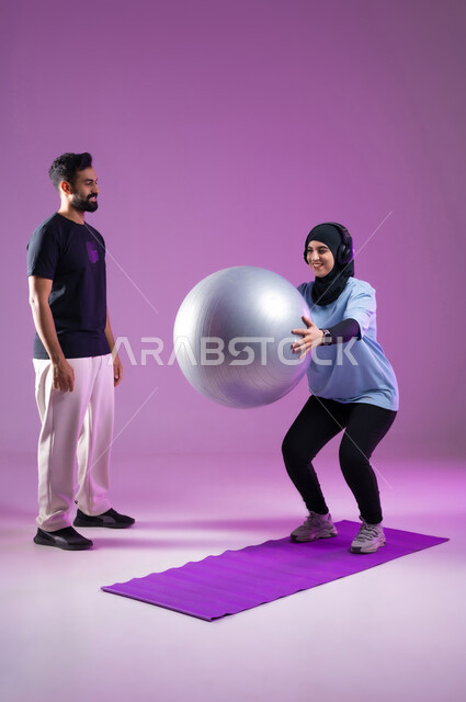 Using a Pilates ball to perform a squat movement, a portrait of a young Saudi Gulf Arab man and a female trainer wearing a headphone while exercising, a healthy lifestyle, the concept of agility and fitness, activities and sports to strengthen the muscles, maintaining physical balance, purple background