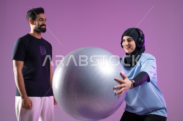 استخدام كرة البيلاتس للقيام بحركة السكوات، بورتريه لشاب ومدربة عربية خليجية سعودية ترتدي سماعه الهيدفون أثناء ممارسة الرياضة ،اسلوب حياة صحي، مفهوم الرشاقة واللياقة البدنية، انشطة ورياضات لتقوية العضلات,  المحافظة على التوازن الجسدي, خلفية بنفسجية