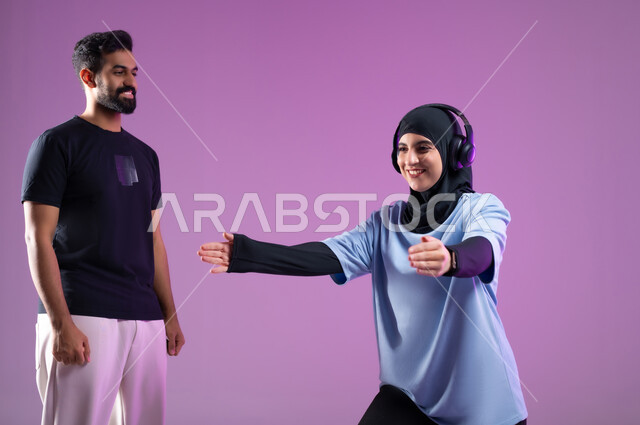 طريقة النزول الصحيحة لحركة السكوات، بورتريه لشاب ومدربة  عربية خليجية سعودية ترتدي سماعة الهيدفون أثناء ممارسة الرياضة ،اسلوب حياة صحي، مفهوم الرشاقة واللياقة البدنية، انشطة ورياضات لتقوية العضلات,  المحافظة على التوازن الجسدي, خلفية بنفسجية