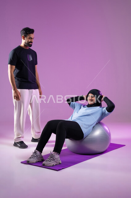 استخدام كرة البيلاتس لممارسة التمارين الرياضية، بورتريه لشاب وامرأة عربية خليجية سعودية ترتدي سماعه الهيدفون أثناء ممارسة الرياضة ،اسلوب حياة صحي، مفهوم الرشاقة واللياقة البدنية، انشطة ورياضات لتقوية العضلات,  المحافظة على التوازن الجسدي, خلفية بنفسجية