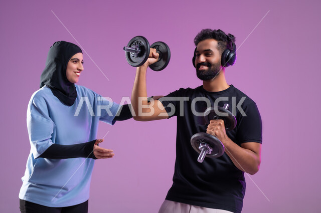 القيام بالاحماء باستخدام ادوات رفع الاوزان، بورتريه لمدربة وشاب عربي خليجي سعودي يرتدي سماعه الهيدفون أثناء ممارسة الرياضة ،اسلوب حياة صحي، مفهوم الرشاقة واللياقة البدنية، انشطة ورياضات لتقوية العضلات,  المحافظة على التوازن الجسدي, خلفية بنفسجية